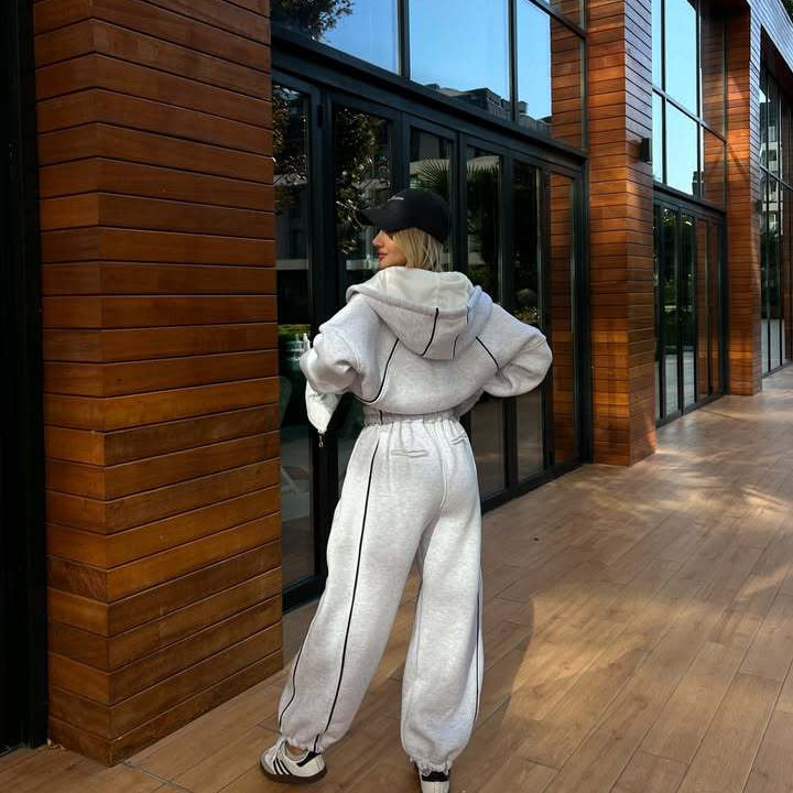 Back view of a model in a light gray oversized hoodie and matching joggers, highlighting the continuous black piping detail running down the sleeves and pants.