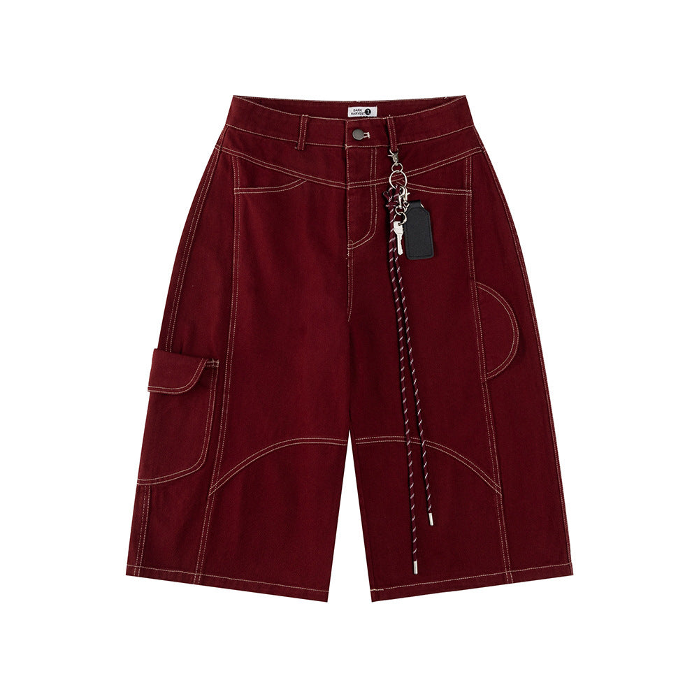 Front flat lay view of relaxed-fit long shorts in a deep burgundy denim, featuring sharp contrast topstitching, asymmetrical utility paneling, a side cargo pocket, and a detachable lanyard with key ring and leather fob accents.