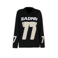 Front studio shot of a black oversized women's V-neck football jersey hoodie featuring a bold cream-colored "BADNM 77" graphic printed on the chest and numbers on the sleeves.